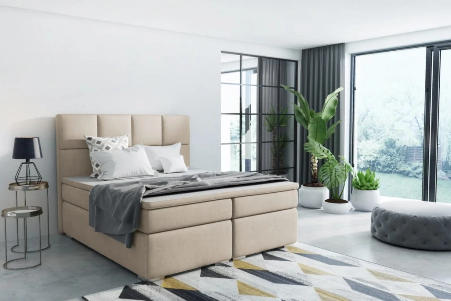Beiges Boxspringbett in hellem Interieur mit Teppich und Zimmerpflanzen.