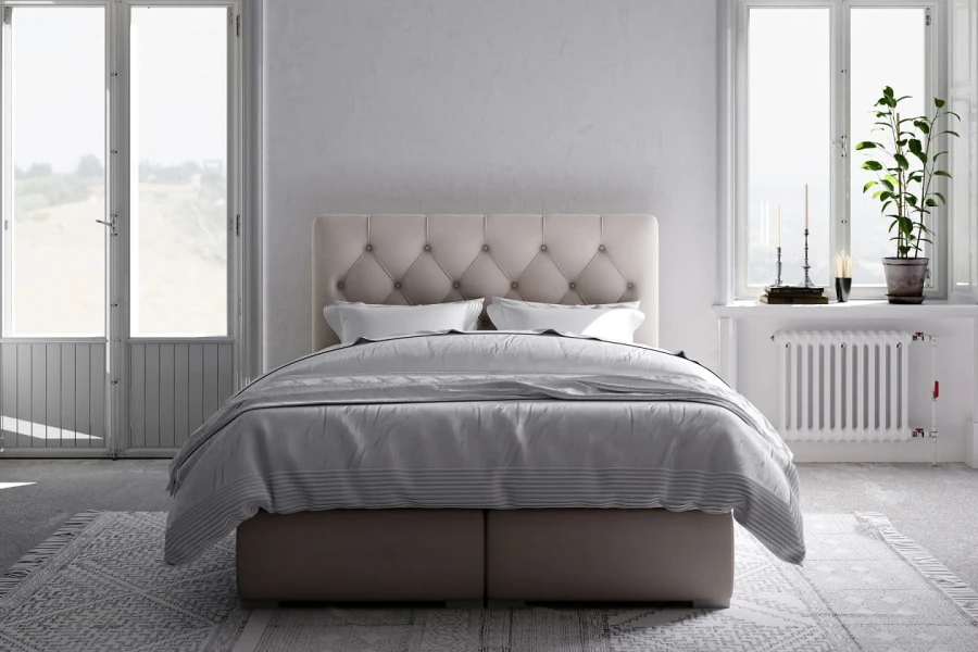 Beiges Boxspringbett mit gestepptem Kopfteil in hellem Schlafzimmer vor einem Fenster.
