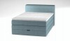 Hellblaues Boxspringbett mit gepolstertem Kopfteil und weißem Topper, schräge Frontansicht.