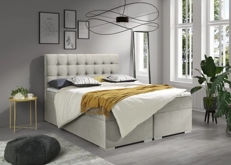Beiges Boxspringbett mit gelber Decke, dekorativer Pflanze und moderner Deckenleuchte.