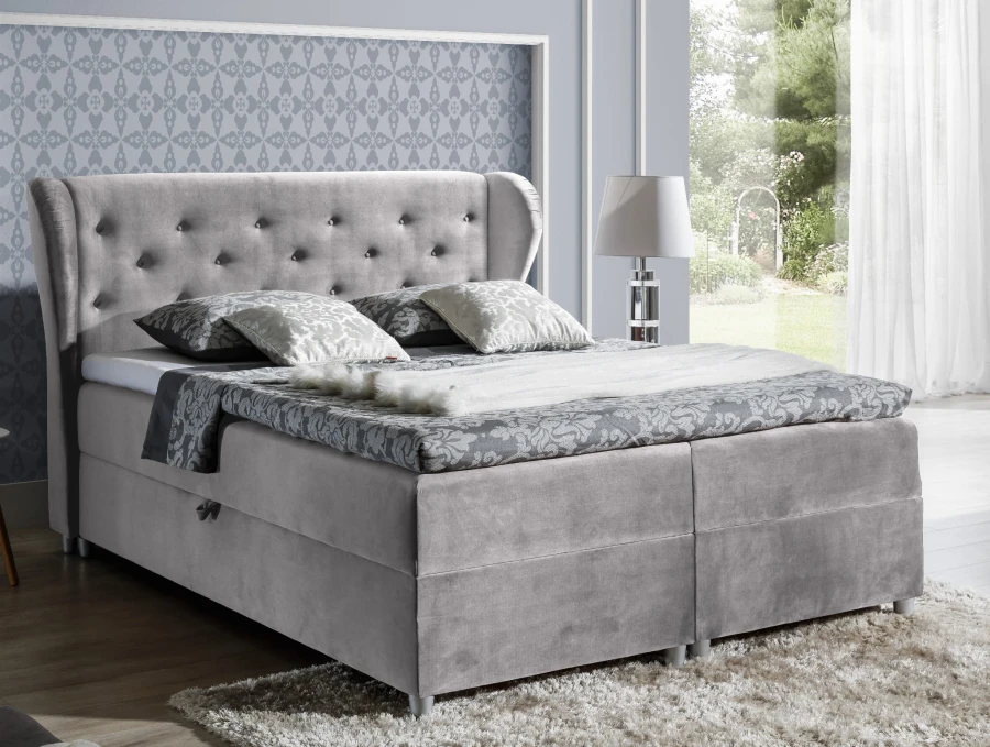 Hellgraues Boxspringbett mit Ohrensessel-Kopfteil und Samtbezug in einem hellen Schlafzimmer-Ambiente.