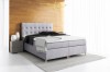 Hellgraues Boxspringbett mit hohem Kopfteil vor Holzwand.
