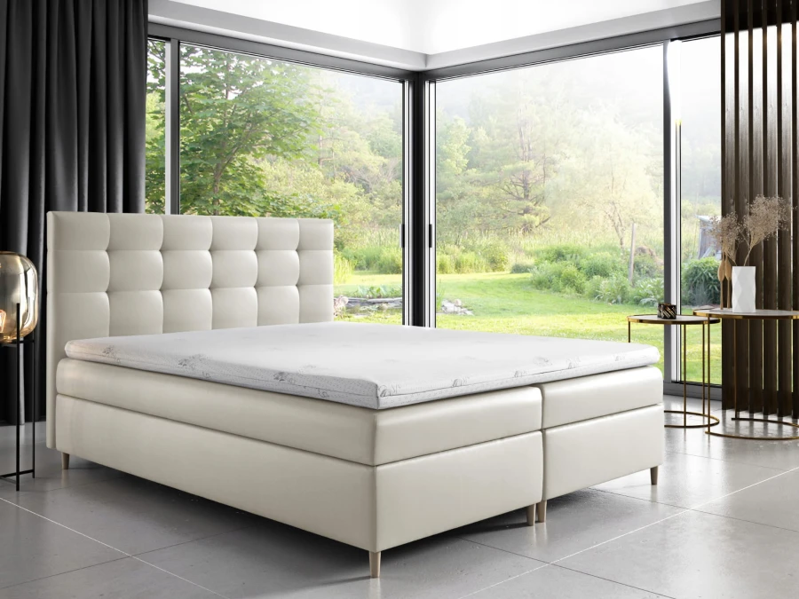 Helles Boxspringbett mit quadratisch gestepptem Kopfteil vor einem großen Panoramafenster.