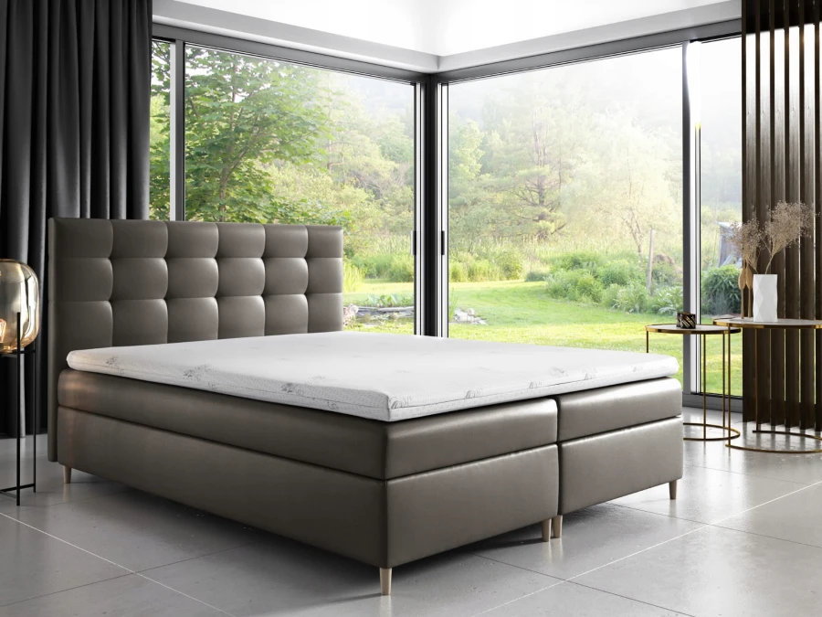 Modernes graues Boxspringbett vor großem Fenster mit Blick ins Grüne.