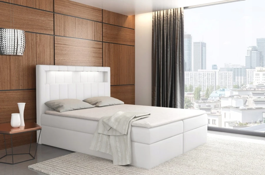 Weißes Boxspringbett mit beleuchtetem Kopfteil vor einer hölzernen Wand.