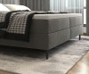 Nahaufnahme eines grauen Boxspringbettes mit weißem Topper und stilvoller Bettwäsche im Schlafzimmer.
