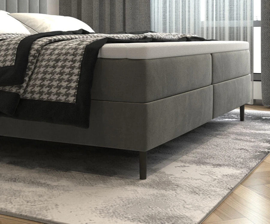 Nahaufnahme eines grauen Boxspringbettes mit weißem Topper und stilvoller Bettwäsche im Schlafzimmer.