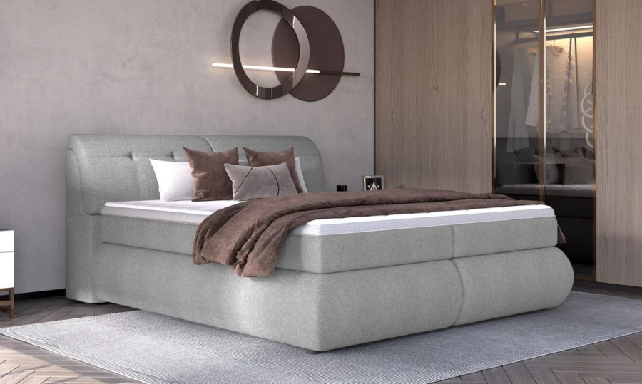Modernes graues Boxspringbett mit gepolstertem Kopfteil und brauner Decke in hellem Schlafzimmer.
