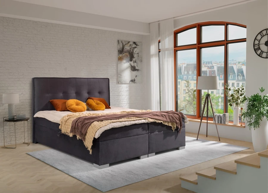 Dunkelgraues Boxspringbett im Schlafzimmer mit großem Fenster und eleganter Einrichtung.