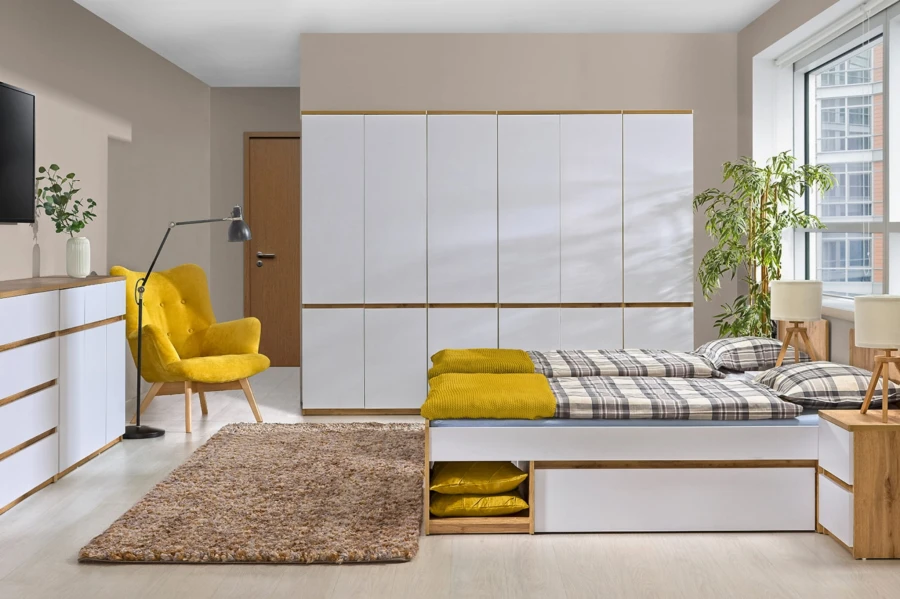 Modernes Schlafzimmer-Set: Weißer Kleiderschrank, Bett mit Stauraum und gelber Sessel vor beiger Wand.