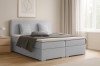 Gezellig boxspringbed in lichtgrijs met kussens en deken in een lichte slaapkamer.