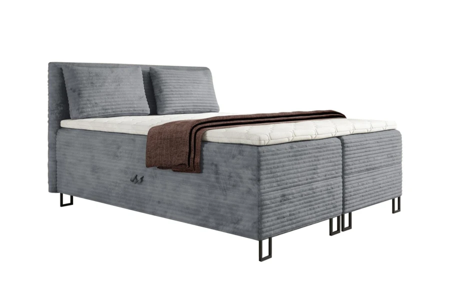 Graues Boxspringbett mit geripptem Stoff, weißem Topper und brauner Decke vor weißem Hintergrund.
