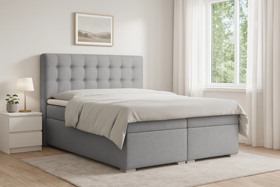 Graues Boxspringbett Claudia im hellen Schlafzimmer mit Fenster, weißem Nachttisch und Teppich.