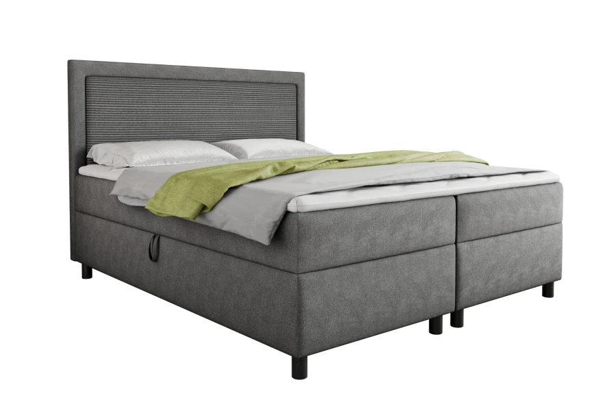 Graues Boxspringbett mit geriffeltem Kopfteil und grüner Tagesdecke, freigestellt.