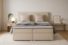 Frontansicht des beigen Boxspringbetts im Schlafzimmer mit Teppich, Nachttisch und Wanddekoration.