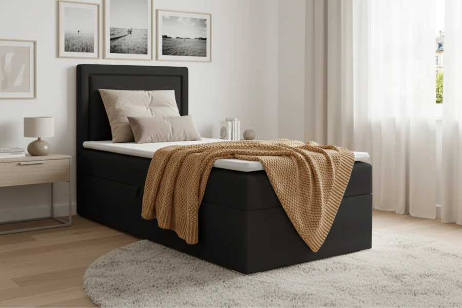 Zwart boxspringbed met opbergruimte, witte topper en gele deken op een lichte houten vloer.