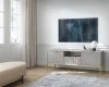 Modernes TV-Lowboard in Hellgrau mit goldenem Gestell und Rillenstruktur unter einem Fernseher.