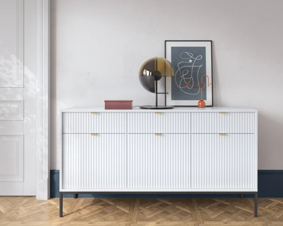 Weißes Sideboard mit geriffelter Front, goldener Lampe und Bild auf Parkettboden.