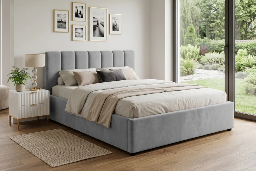 Eingerichtetes Schlafzimmer mit grauem Polsterbett, Nachttisch und Blick in den grünen Garten.
